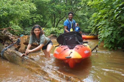 A young woman and man navigate kayaks around logs in a shallow creek surrounded by lush greenery
