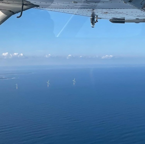 small wind turbines in the middle of of the ocean are seen from the air, with the bottom of a plane above