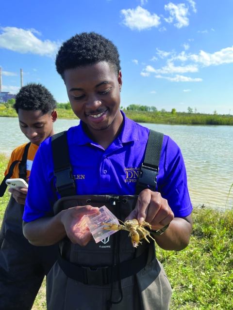A young man in waders smiles and holds a crayfish in front of a shallow freshwater lake