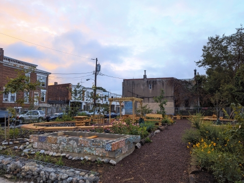 A garden with wooden and stone built features and green foliage in the foreground with buildings of a city block in the background