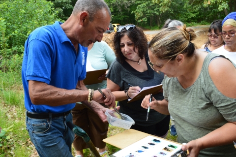 a man holds a tupperware and hand tool up for a group of women to look at. The women hold up clipboards and pens with paper featuring a variety of insect species.