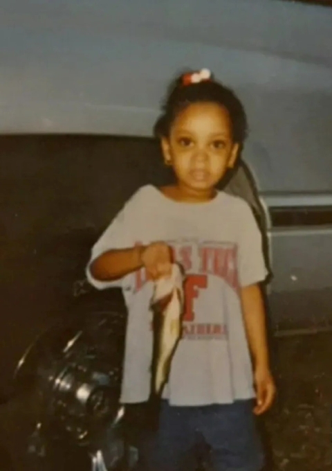 A little girl wearing a t-shirt holds a small fish while standing in front of a car