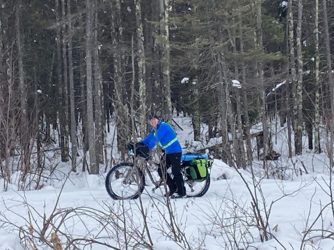 A man in a blue jacket pushes his bike along a snow-covered trail in the woods.