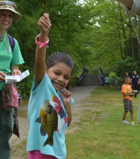 a young girl smiles while holding a fishing line to show off a fish she caught. She's standing in an urban park. An adult woman, children and law enforcement are seen standing in the background. 