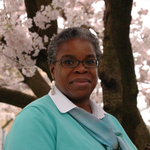 A woman wearing glasses and a blue sweater smiles while standing under a blooming cherry blossom tree