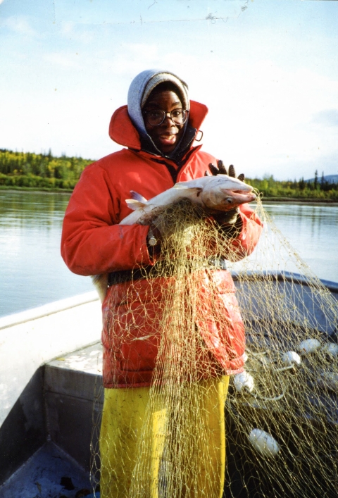A woman stands in a boat smiling and holding a fish and a net. She wears a heavy orange coat and yellow rain pants.