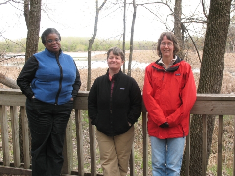 Three women smile and stand along a rail in a wooded area. 