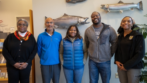 Five people pose for an office photo with adult fish sculptures mounted on the wall behind them.