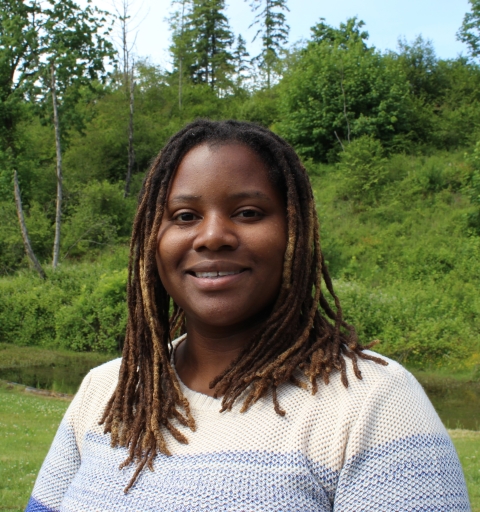 A woman smiles at the camera. Behind her are green trees and a small body of water.