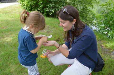 In an outdoor green space, a young woman holds up a small insect species in her hand for a young girl to inspect