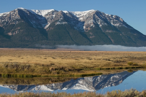 Snow-capped mountains in the background rise from the landscape and provide a reflection in a pond in the foreground.