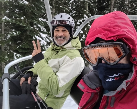 Two men sit on a ski lift chair, one smiles wearing a green and white jacket, the other is wearing a red jacket and goggles and a face covering. Green trees with snow are in the background/