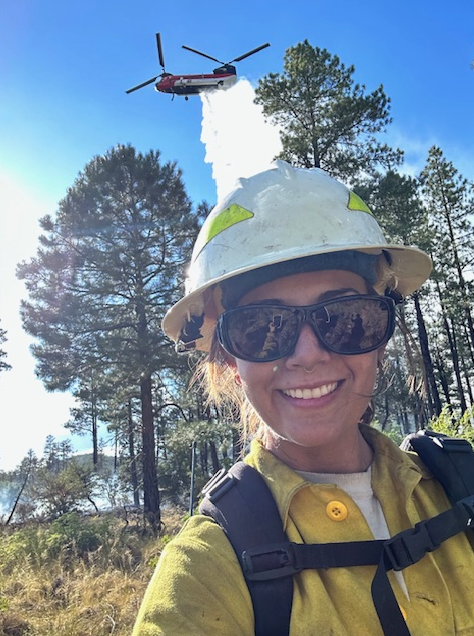 A firefighter takes a selfie during suppression operations with a helicopter dropping water behind her