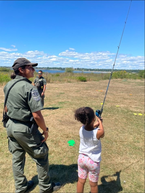Officer smiling as a young girl casts a fishing rod on a green lawn outside. 