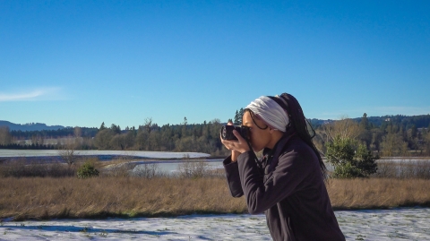 A woman looks through a camera facing left and takes a photo. There is snow and dry grassy areas behind her with a blue sky.
