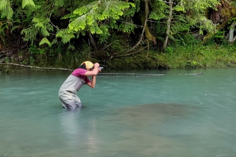 A woman wearing waders stands knee deep in water while taking a photo