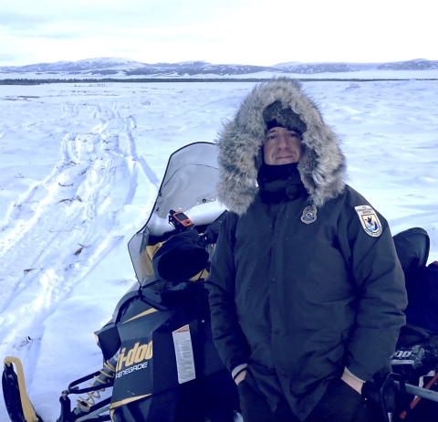 A man dressed in a Fish and Wildlife Service jacket and fur-lined hood stands along a snowmobile on a vast field of snow.