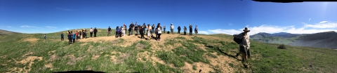 A wide view of about 30 students on a grass hilltop with blue sky background