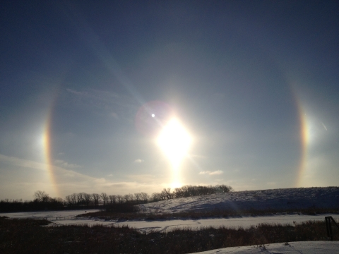 A bright sunrise flanked by two rainbow sundogs which extend up into fading arcs over Mallard Marsh in winter