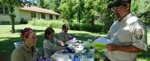 People sit at a table in brown uniforms outdoors dissect fish while another person stands with a clipboard