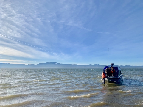 A boat with three people wearing orange life vests floats on a large river with choppy waves. Mountains and a blue sky with clouds visible in the background.