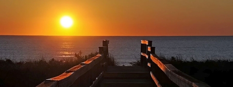 In the distance, the sun rising over the Atlantic Ocean amid an orange-yellow glow. In the foreground a boardwalk trail leading to the ocean.