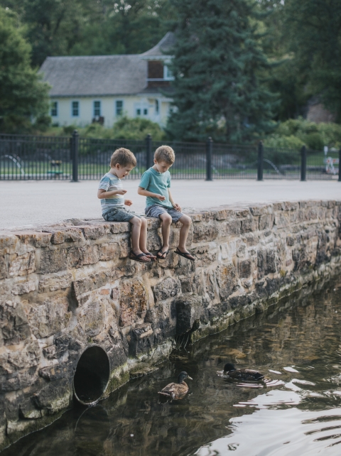 Two young boys sit on the edge of a stone walled pond and feed the trout and ducks. The historic 1899 hatchery building is visible in the background.