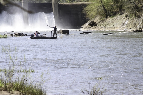 Boat in a river just downstream of a dam. Two people in the boat, one operating the motor while the other stands in the bow with a net