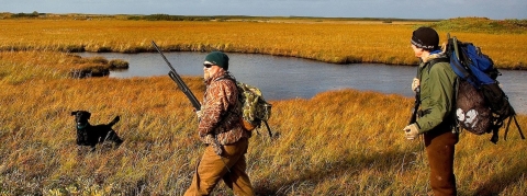 Two men carrying guns and backpacks walk across grassland next to a body of water with their hunting dog