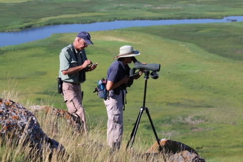 Volunteers Chuck and Betty Mulcahy, wearing hats and hiking clothes, survey trumpeter swans and cygnets weekly from atop a butte at National Elk Refuge in Wyoming.