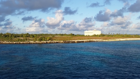 An abandoned building sits on Johnston. The blue ocean water meets the rocky shoreline of the island that is covered in green trees and grass. Blue skies light up the background. 