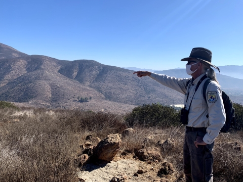 Biologist John Martin pointing at something by shrubs. Rolling mountains in the background. 