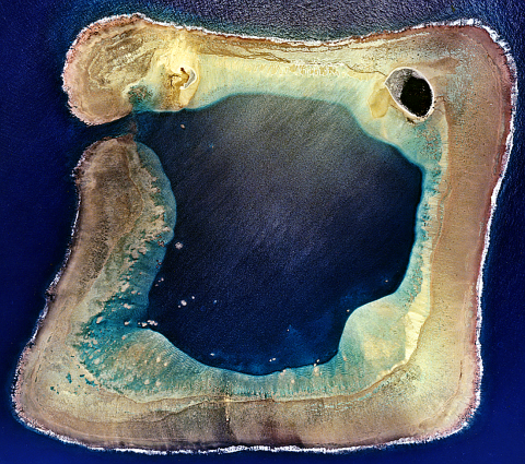 A satellite view of Wake Atoll. It is shaped like a square with an entrance to the sea to the left that opens up to a large bay. 