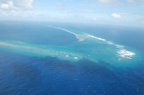 An aeriol view of Kingman Reef. Light blue outlines the shallow areas of the reef while the deeper dark blues of the ocean surround the area. 