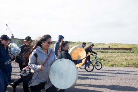 Woman with white skin drum leads group of people walking down a road. Kids on bikes flank her.