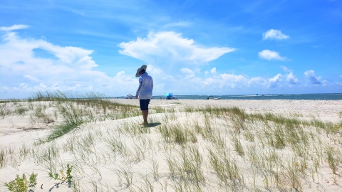Kid with a hat stands on a dune with a sliver of ocean and a blue cloudy sky in the background..