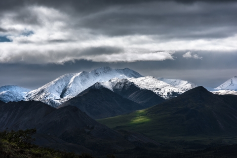 Snow-capped mountains loom over a green valley, all under stomy clouds