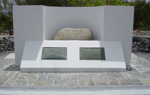 A memorial on Wake Atoll. A round stone with Japanese words inscribe on it sits on a white alter. Below on the stone are two engraved plaques with Japanese words on them. 