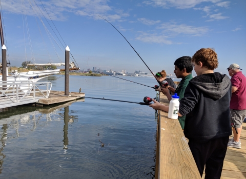 Three students fish from pier at Masonville Cove with Baltimore skyline in background