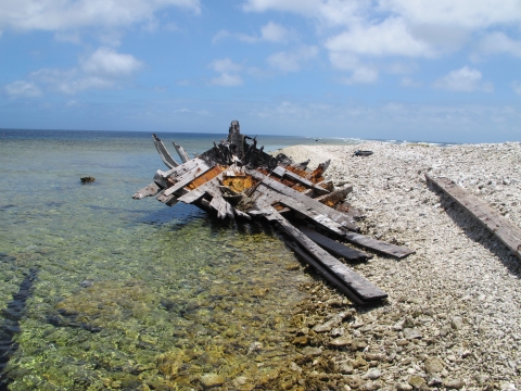 The wooden wreck of a ship lays beached on the shores of Kingman. Water is to its left with rocks to its right. 