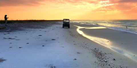 A bright orange sunrise looking down a coastal beach with a silhouette recording data from the dunes, and a golf cart midway between the silhouette and the surf