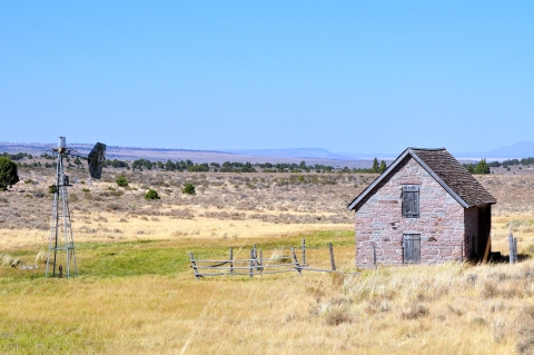 An old stone A-framed building in a high desert landscape with an old mill next to it