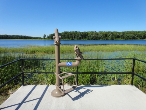 View of a overlook platform on the edge of a wetland that has two spotting scopes that are at two different levels, with the lower one being accessible and having steps for children. It overlooks a wetland scene with aquatic grasses and lily pads in the foreground and open water beyond, with the far shore lined with trees.