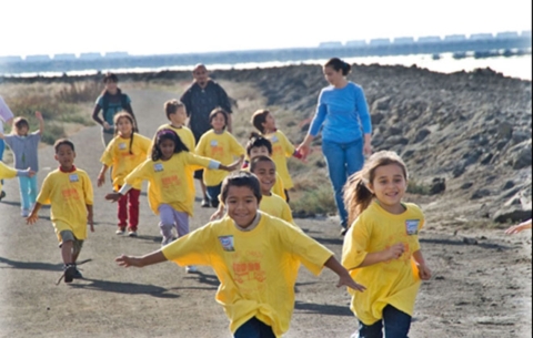 Children in matching over-size yellow tee shirts run on a dirt trail, with three adults behind them