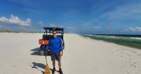 Under a partly sunny sky, facing the camera is a man wearing a blue shirt standing on a white sand beach in front of an off road beach club car.