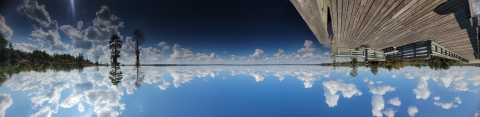 Image of clouds reflecting over a lake with cypress trees on the edge and a wooden pier