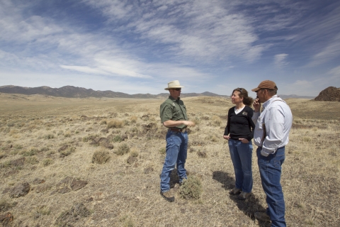 Two men in hats meet with a woman on a piece of arid-looking land.