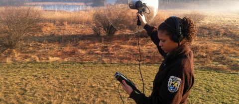 A woman in a U.S. Fish and Wildlife Service uniform and wearing headphones holding a sound cone in the air in an open field with a body of water in the distance