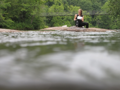 Woman sitting on a rock in a river, writing on a clipboard