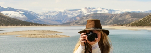 A woman looks through a camera at the person taking her picture. Behind her are snow-covered mountains and a lake